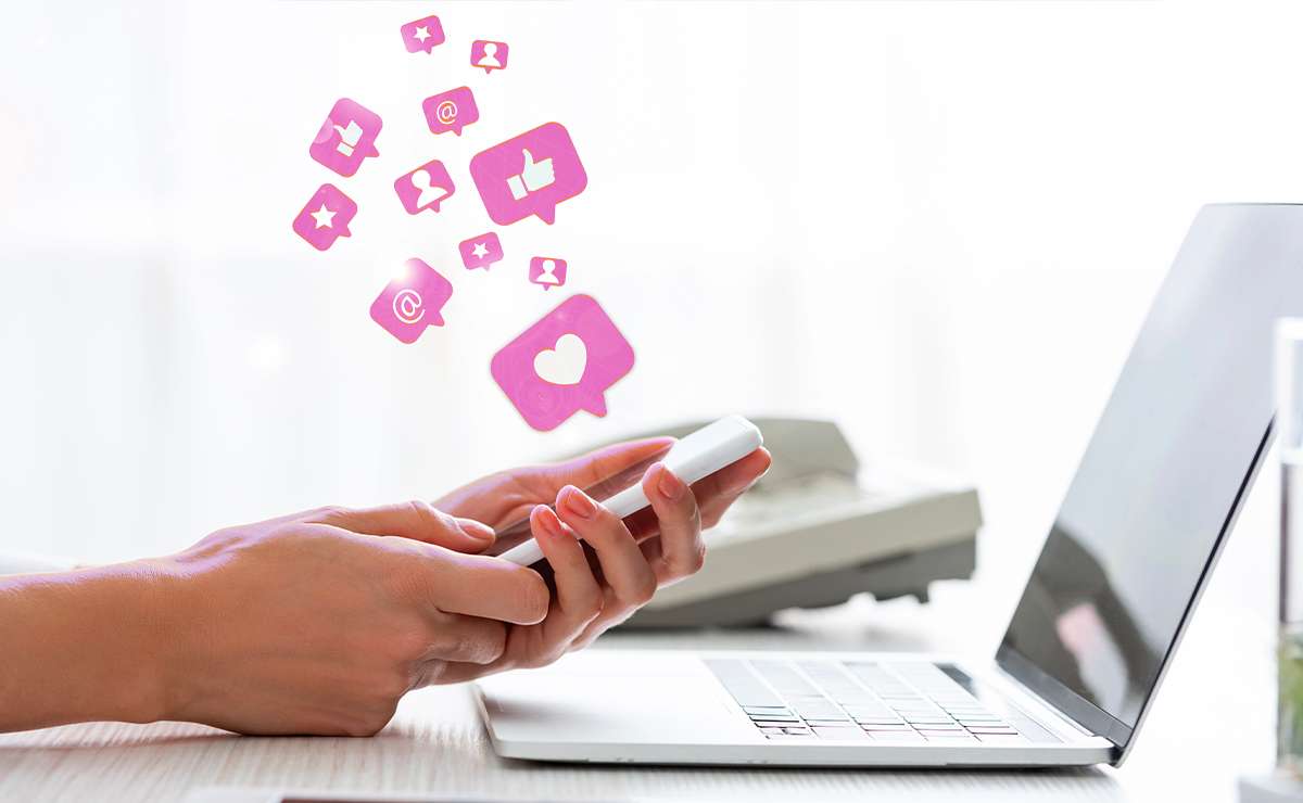 Persona che utilizza uno smartphone con icone di interazione social, accanto a un computer portatile.